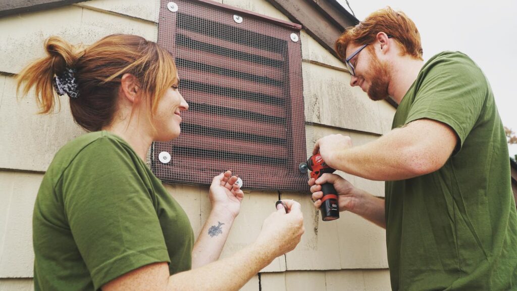 A man uses a drill to secure wire mesh to a vent on a house.